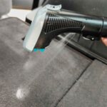 Poignée d'un injecteur-extracteur en action, pulvérisant de l'eau et du produit pour nettoyer un siège de voiture. (c) Labo Maison
